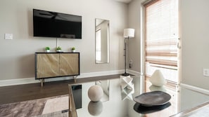 Uma sala de estar moderna com TV de tela plana, um espelho grande, luminária de chão e mesa de centro de vidro com objetos decorativos.