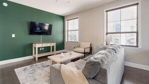 A living room with a green accent wall, a flat-screen TV, a sofa, a coffee table, and a small table with a lamp. - Landing | Amazing 2BD, Gym, Courtyard (San Antonio)
