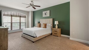 Quarto com cama, mesa de cabeceira, ventilador de teto, janela com persianas e um quadro na parede.