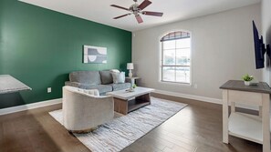 A living room with a green wall, a grey sofa, a coffee table, a white armchair, a small table with a lamp, and a window with blinds. - Landing | Amazing 1BD, Courtyard, Gym (San Antonio)
