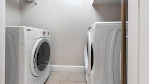 A laundry room with two front-loading washing machines, a white tiled floor, and a white wall. - Landing | Modern 2BD, Gym, Courtyard (San Antonio)