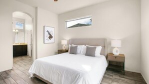 A bedroom with a large bed, two bedside tables with lamps, a window with a view, and a bathroom visible through an archway. - Landing | Stunning 1BD, Courtyard, Gym (San Antonio)
