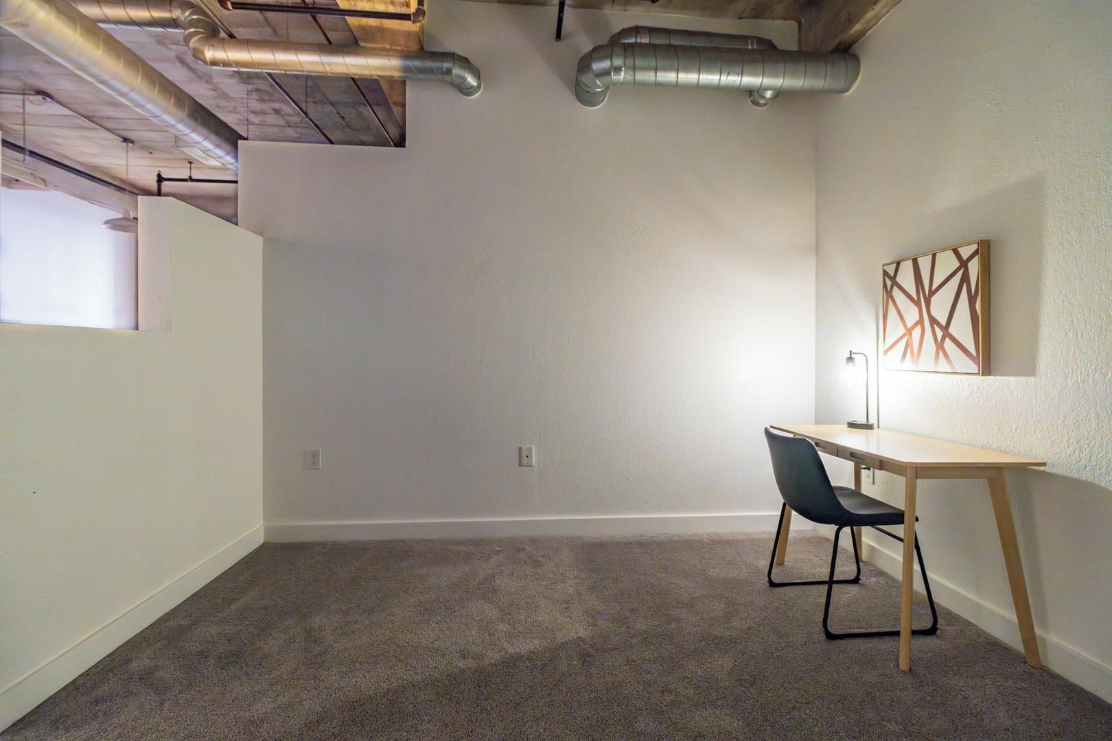 A room with a carpeted floor, a desk with a chair, a wall-mounted lamp, and a framed artwork.