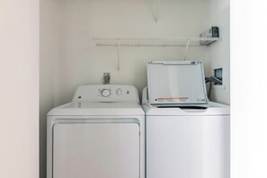 A laundry room with two white washing machines, a metal shelf above, and a power outlet on the wall. - Landing | Chic 1BD, Pool, Gym (Kansas City)