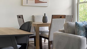 Et spiseområde med et træbord og stole, en vase på bordet og et vindue med udsigt til grønne omgivelser.
