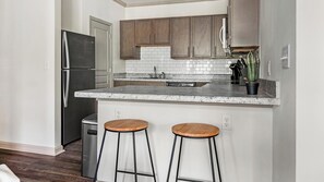 A kitchen with a stainless steel refrigerator, wooden cabinets, a granite countertop, and two bar stools.
