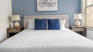 A neatly made bed with white bedding, two blue pillows, and a grey headboard. There are two bedside tables with lamps and a framed artwork above the bed.