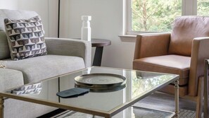 Un salon moderne avec une table basse en verre, un canapé gris, un fauteuil en cuir marron et une vue sur les arbres par la fenêtre.