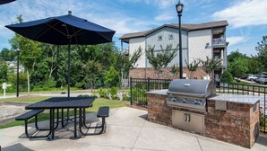 En udendørs terrasse med grill og siddeplads, omgivet af en murstensmur og en bygning med balkon.