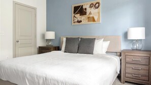 A neatly made bed with a white duvet, a headboard with two pillows, a bedside table with a lamp, and a wall-mounted abstract artwork. - Landing | Beautiful 1BD, Gym (Gainesville)