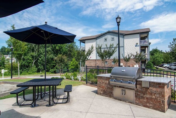 An outdoor patio with a grill and seating area, surrounded by a brick wall and a building with a balcony. - Landing | Chic 1BD, Gym (Gainesville)