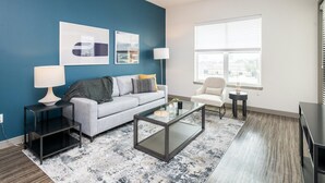 Een moderne woonkamer met een grijze bank, een glazen salontafel, een bijzettafeltje met een lamp, een witte fauteuil en een groot raam met jaloezieën.