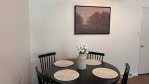 Ruokailutilassa on pyöreä pöytä, neljä tuolia, maljakko kukkineen ja seinällä oleva kehystetty kuva.