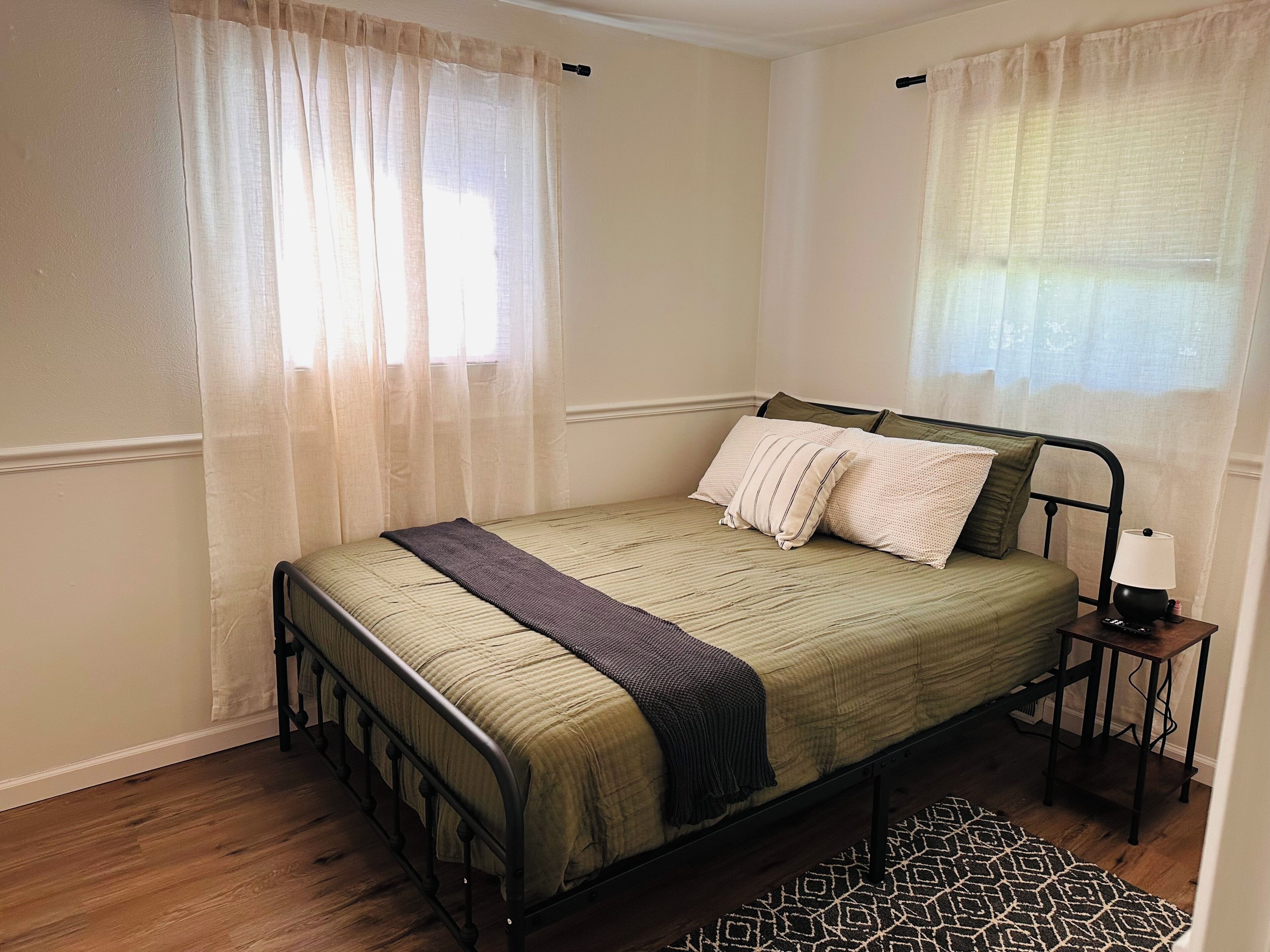A single bed with a metal frame, a bedside table with a lamp, and a patterned rug on a wooden floor.