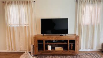 Una sala de estar con un televisor de pantalla plana sobre un mueble de madera, dos ventanas con cortinas traslúcidas, una alfombra estampada y un sofá con cojines a rayas.