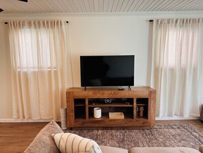 Una sala de estar con un televisor de pantalla plana sobre un mueble de madera, dos ventanas con cortinas traslúcidas, una alfombra estampada y un sofá con cojines a rayas.