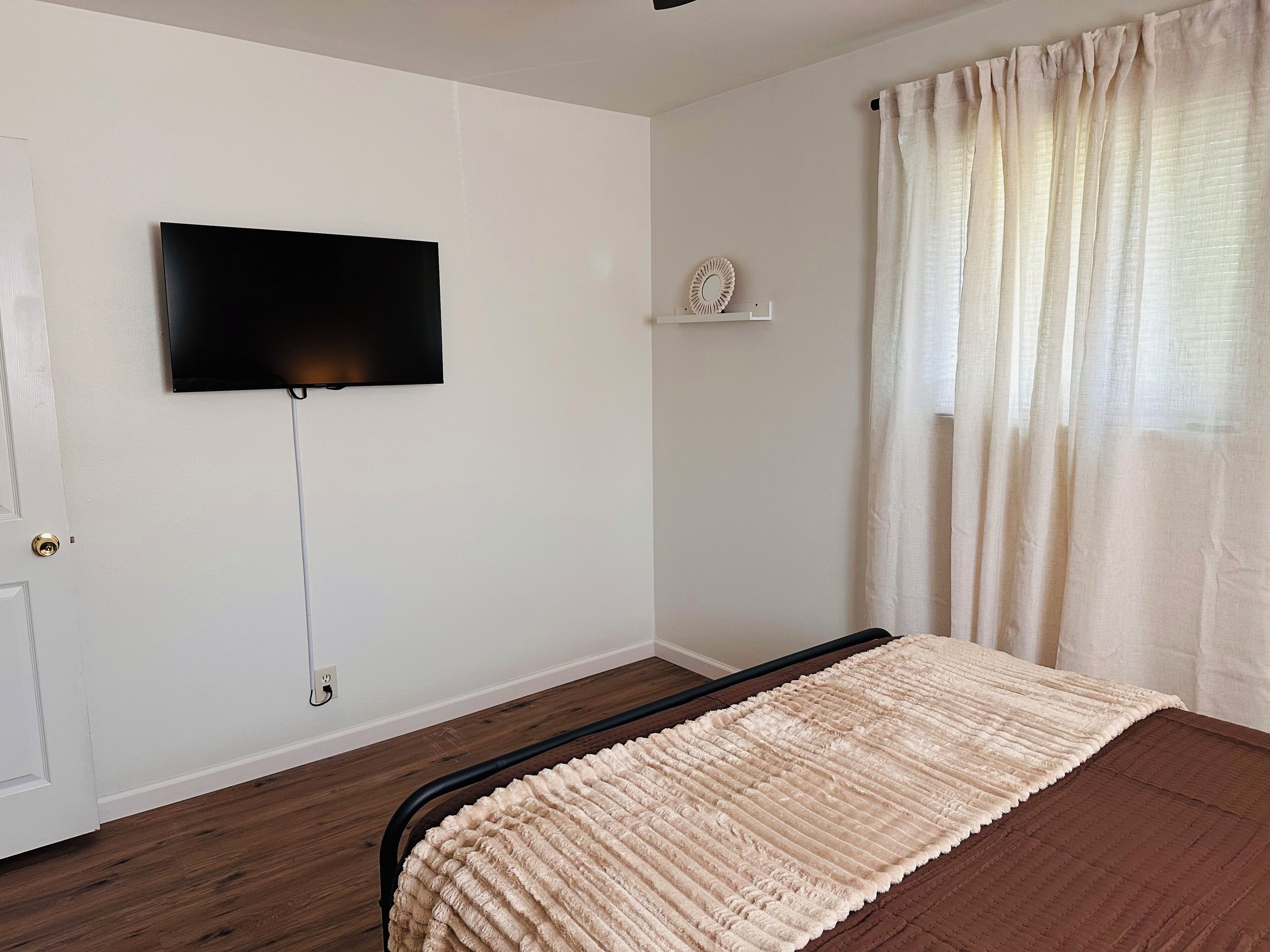 A bedroom with a bed, a television mounted on the wall, a window with curtains, and a door.