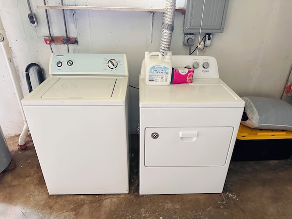 Two white washing machines in a room with a concrete floor, a metal vent, and a wooden shelf above.