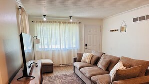 A living room with a brown sofa, a wooden coffee table, and a patterned rug. There is a door and a window with sheer curtains.
