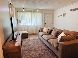 A living room with a brown sofa, a wooden coffee table, and a patterned rug. There is a door and a window with sheer curtains.