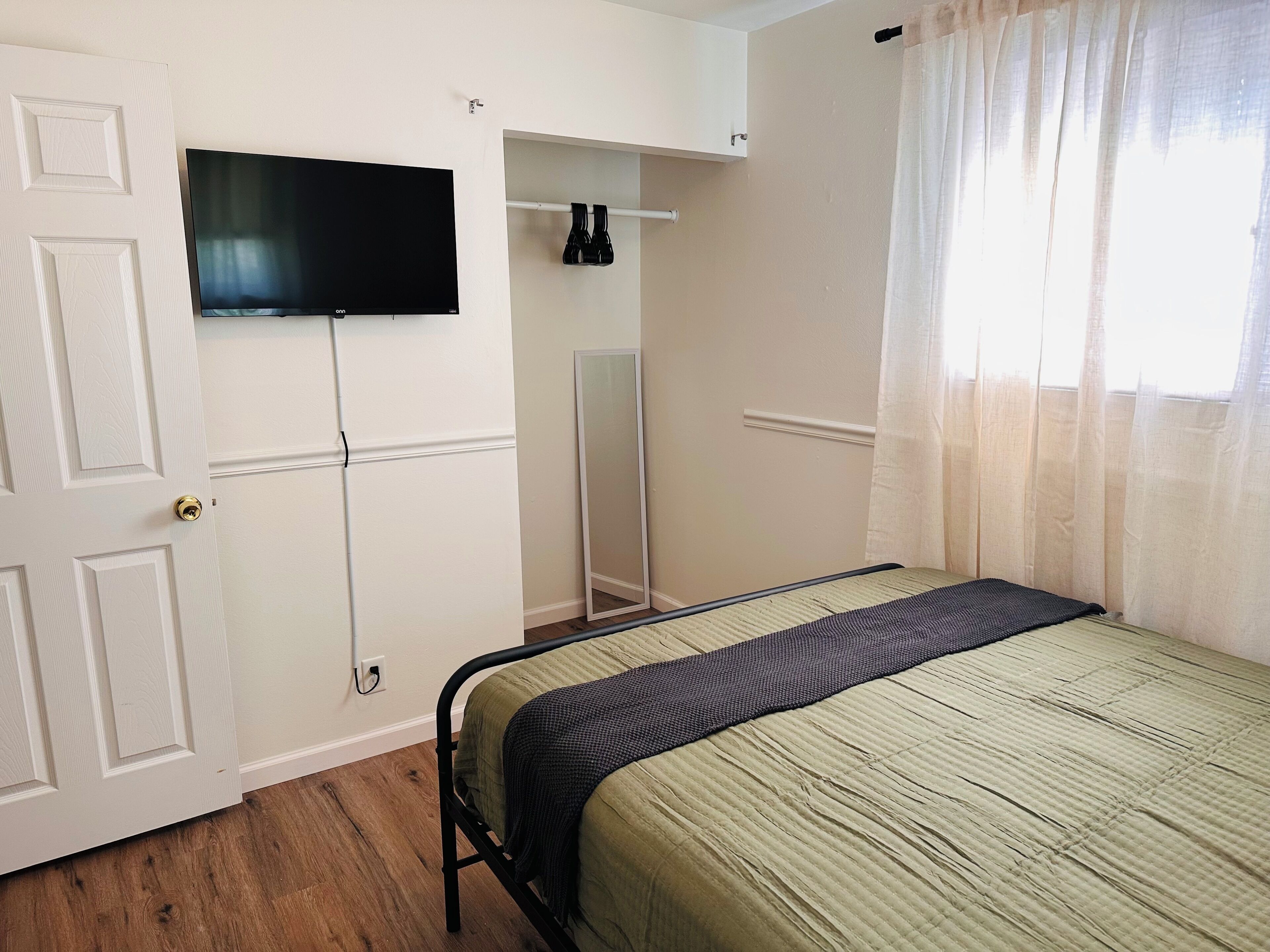 A bedroom with a bed, a television mounted on the wall, a mirror, and a window with curtains.
