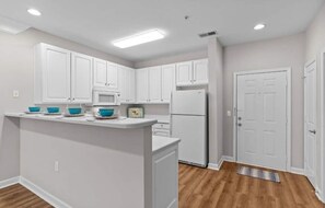 A kitchen with white cabinets, a microwave, a refrigerator, and a small island with blue bowls. - 3-bedroom condo in beautiful Calabash golf course community (Calabash)