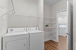 A laundry area with two white washers and dryers, metal shelving units, and a door leading to a kitchen area with white cabinets and blue bowls. - 3-bedroom condo in beautiful Calabash golf course community (Calabash)