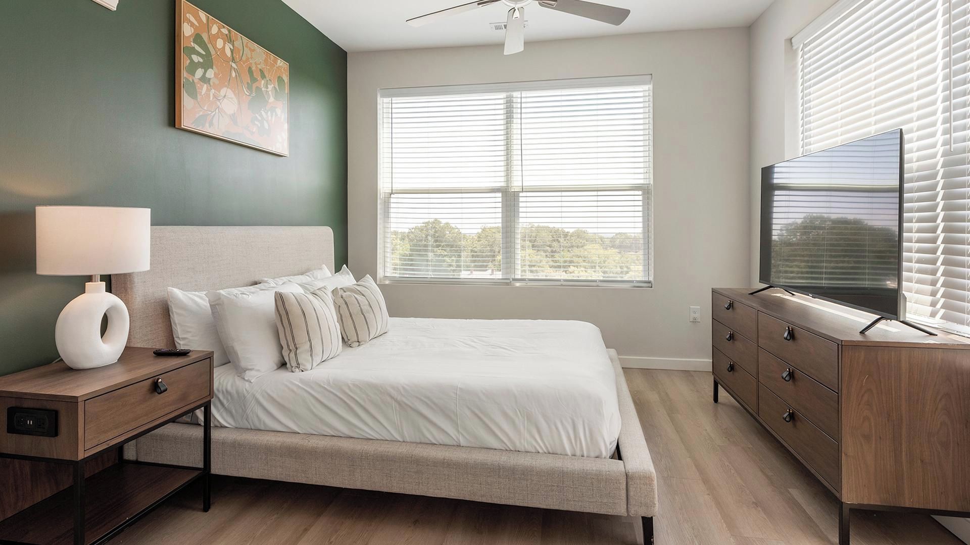 A bedroom with a bed, nightstand, lamp, dresser, TV, and a window with blinds.