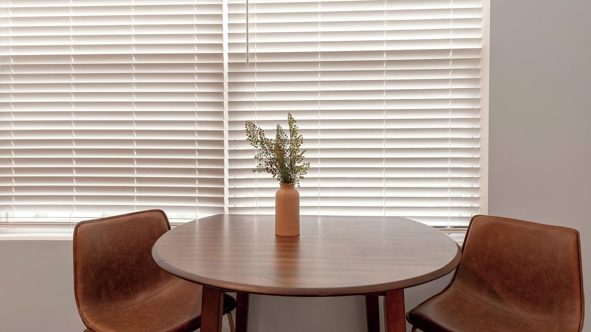 A round table with two chairs, a vase with a plant, and white blinds in the background.