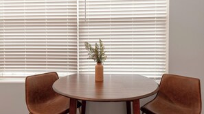 Ein runder Tisch mit zwei Stühlen, einer Vase mit Pflanze und weißen Jalousien im Hintergrund.