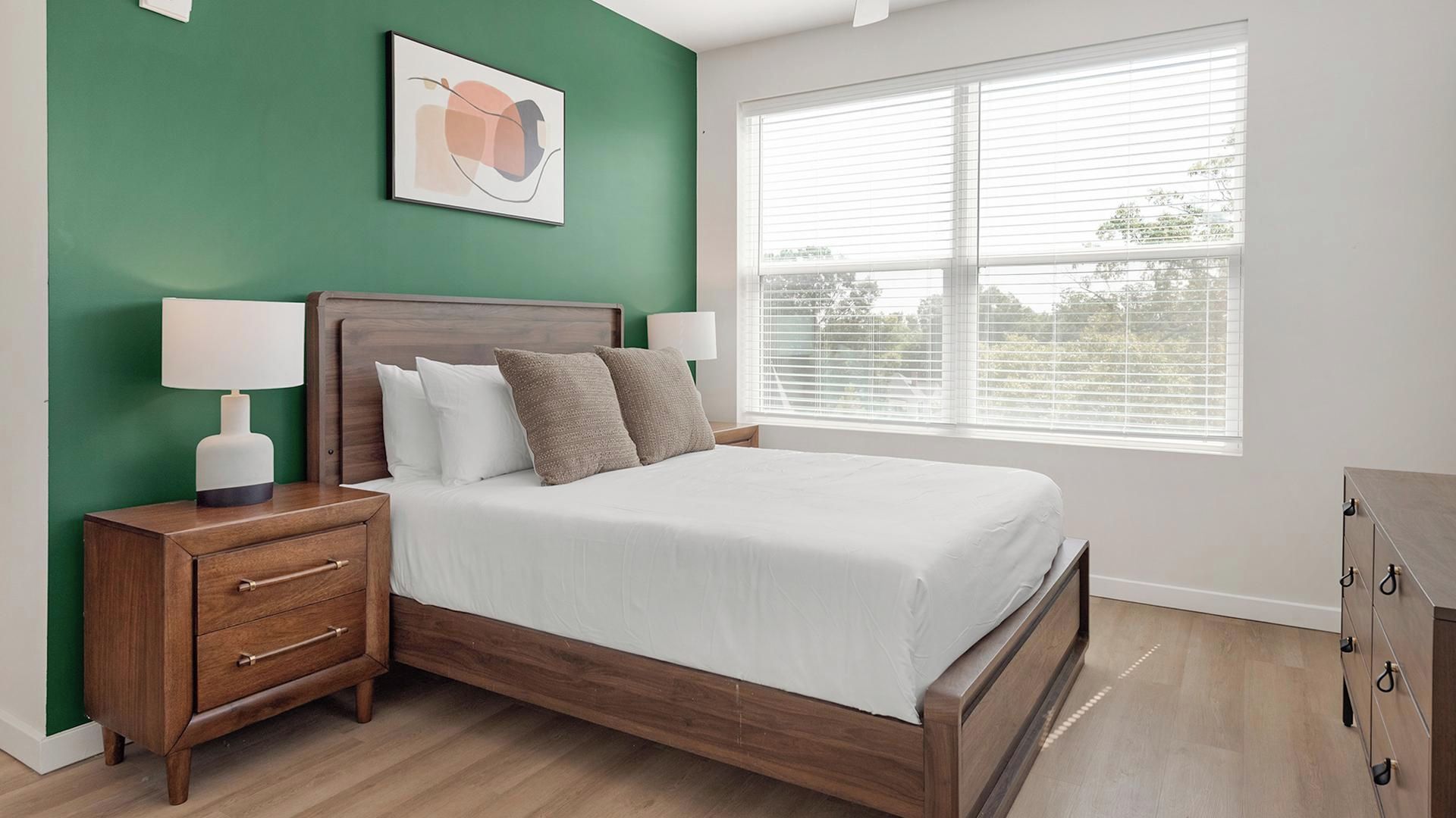 A bedroom with a bed, nightstands, a lamp, a dresser, and a window with blinds.