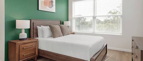 A bedroom with a bed, nightstands, a lamp, a dresser, and a window with blinds.