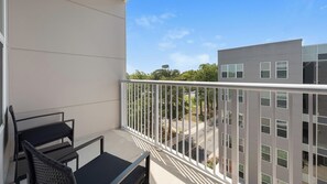A balcony with a white railing, two black chairs, and a view of a modern building and trees. - Landing | Modern 1BD in Kannapolis (Kannapolis)