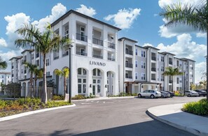 A multi-story building with a name sign 'LIVANO' on the facade, featuring balconies, a parking area with cars, and a clear sky. - Landing | Spectacular 1BD, Pool, Gym (Port Charlotte)