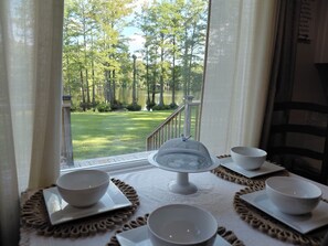A dining table with four bowls and a cake server, set on a tablecloth with a view of a grassy area and trees through a window.