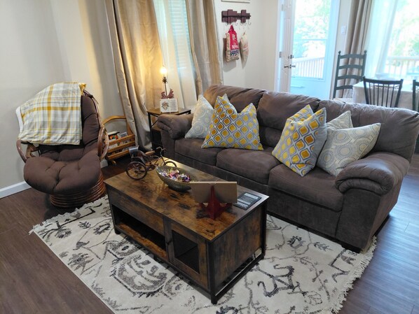 A cozy living room with a brown sofa, patterned cushions, a wooden coffee table with a bowl and a book, a wicker chair, and a patterned rug.