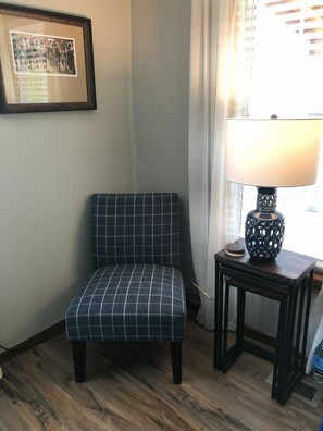 Une pièce avec un fauteuil à carreaux, une table avec une lampe et un tableau encadré au mur.