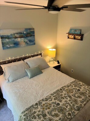 A bedroom with a bed, a nightstand, a lamp, a ceiling fan, a painting, and a sign on the wall. - Charming 3-bedroom cabin with easy lake access, huge back deck and fun extras! (Clarkson)