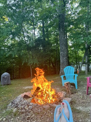 A campfire with a tent and chairs in a wooded area.