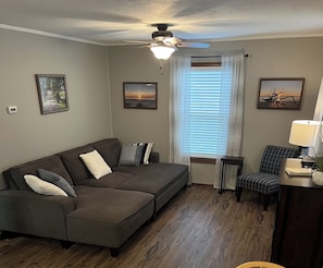 A living room with a sectional sofa, a ceiling fan, and a window with blinds.