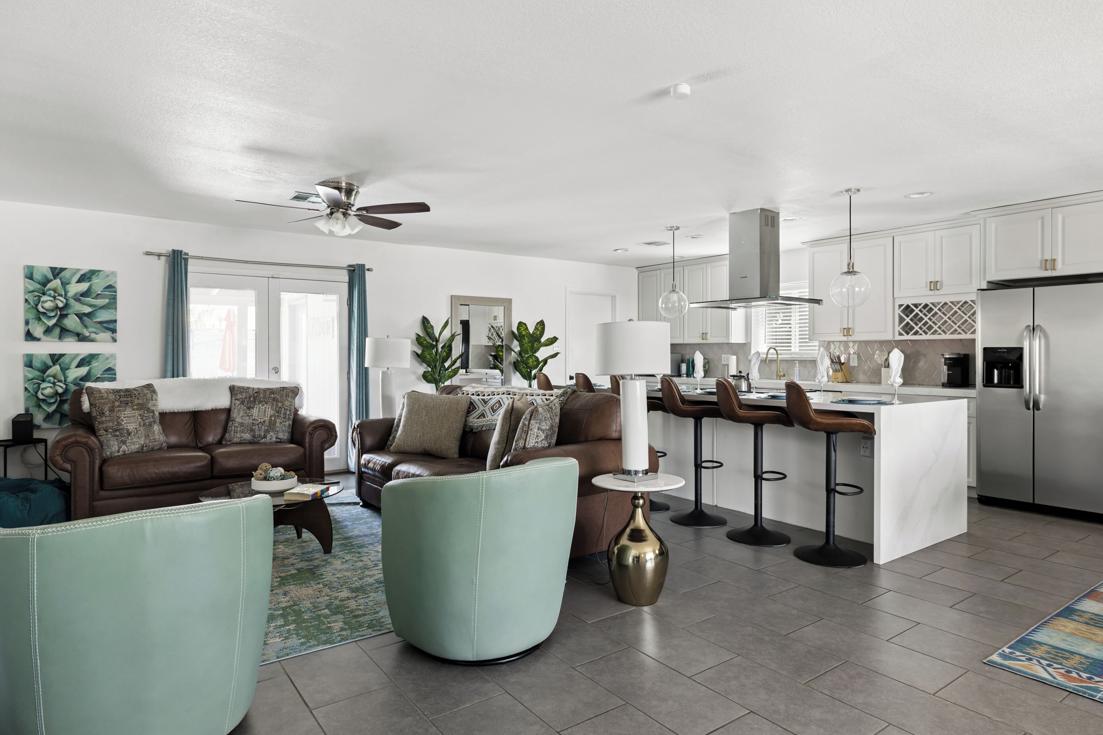A modern living room with a sofa, armchairs, and a coffee table. There is a kitchen area with a refrigerator, stove, and bar stools.