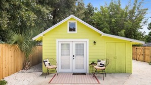 A small, single-story building with a green exterior, a white door, and a small window above the door. There are two chairs outside the building, a small table with a plant, and a red and white rug on the ground. - King Studio · Rainfall Shower · Beach Gear (Bradenton)