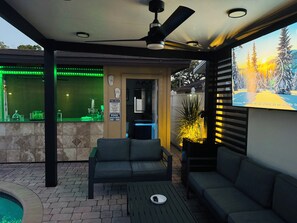 A covered outdoor seating area with a pool, a ceiling fan, and a TV screen displaying a scenic view. - West Tampa Villas # 3 Near Stadium & Airport And Pool (Tampa)
