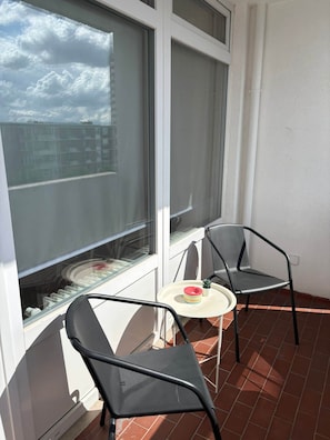 A balcony with two chairs and a small table, a window with a view of buildings, and a white wall.