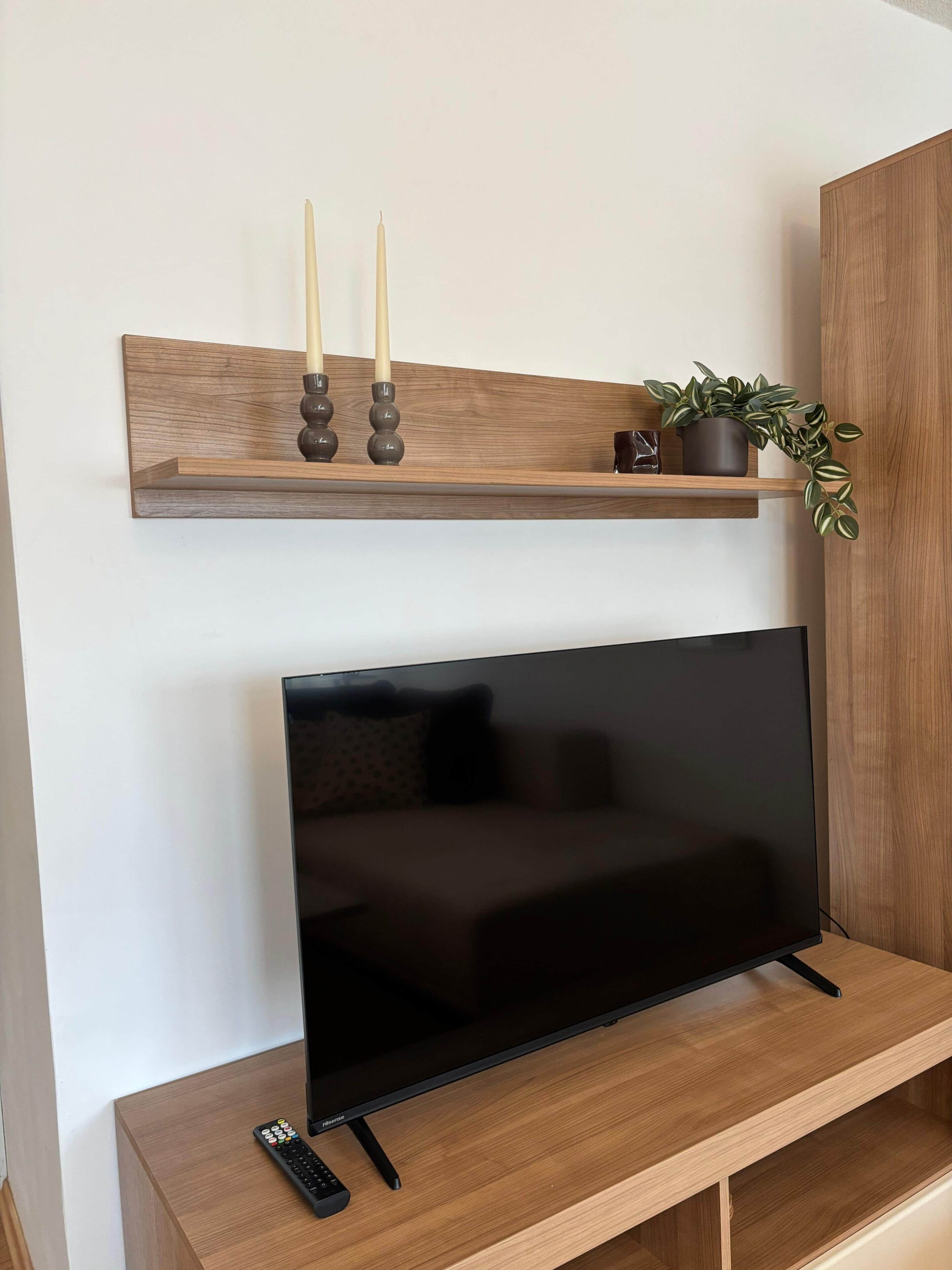 Un téléviseur à écran plat est posé sur un meuble en bois, avec une télécommande à côté. Au-dessus du téléviseur, on aperçoit une étagère en bois ornée de bougies et d’une plante en pot.