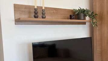 Un téléviseur à écran plat est posé sur un meuble en bois, avec une télécommande à côté. Au-dessus du téléviseur, on aperçoit une étagère en bois ornée de bougies et d’une plante en pot.