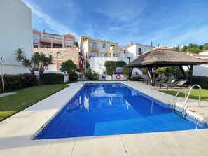 A swimming pool with a clear blue water surface, surrounded by a paved deck, a lifebuoy, and a thatched-roof gazebo.