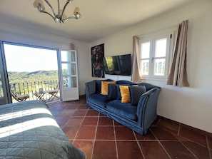 A room with a bed, a blue sofa with yellow cushions, a TV, and a balcony with a view of the countryside.
