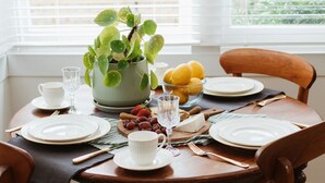 Una mesa de comedor dispuesta con platos, cubiertos, una copa, un cuenco de limones, una planta en maceta y un bol de uvas.