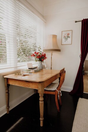 A dining area with a wooden table, a chair, a vase with flowers, a lamp, and a window with blinds. - Whimsy Cottage | Kingston Beach Retreat with Outdoor Bath (Kingston Beach)